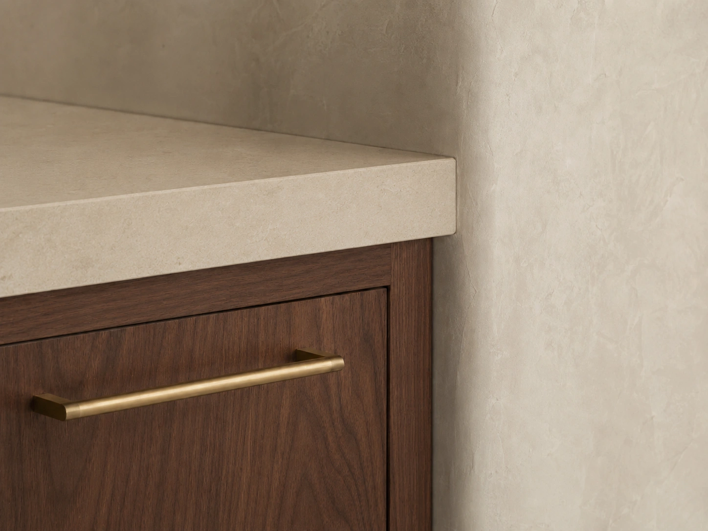 Concept detail image of a cabinet corner in warm walnut with a honed limestone counter edge and a brushed brass bar pull against a soft plaster wall.